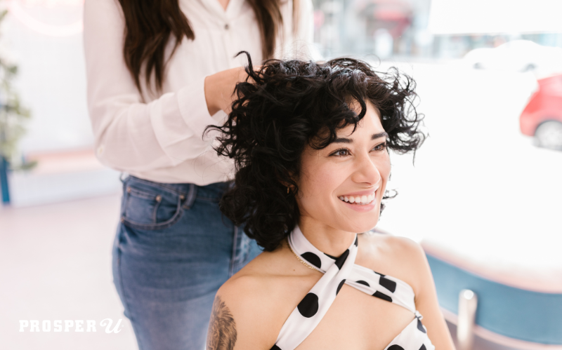 Smiling young woman with short, curly dark hair sits in a hair stylist's chair looking forward in the mirror towards her stylist.